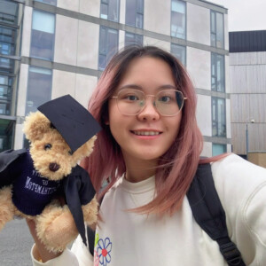 Photograph of a woman standing in front of a building and holding a teddy bear dressed in mortar board and gown.