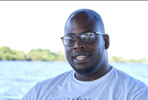 Photograph of the head and shoulders of a man in front of a body of water with trees or bushes in the distance.