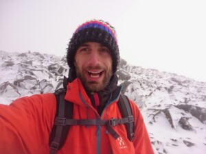 A person in a bright orange jacket and colourful knitted hat with a backpack, standing in front of a snowy mountain landscape.
