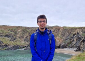 Photograph of a man standing in front of a coastal landscape, with sloping cliffs and a small sandy beach.