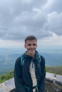 Photograph of a man standing on a high outdoor viewpoint in front of a wide landscape.
