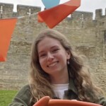 A photograph of the head and shoulders of a woman with a castle wall in the background.