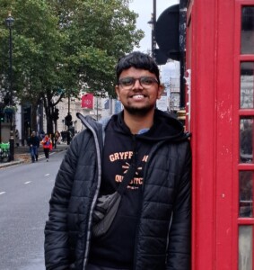 Photograph of a man learning against a traditional red telephone box on an urban street.