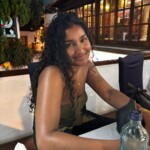 Photograph of a woman seated at a table outside a bar or cafe.