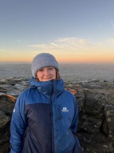 A photograph of the upper body of a woman standing on a rocky outcrop in front of a wide landscape.