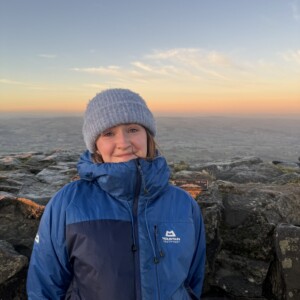 A photograph of the upper body of a woman standing on a rocky outcrop in front of a wide landscape.