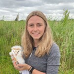 A photograph of a woman holding a baby barn owl and standing in a field.