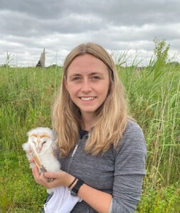A photograph of a woman holding a baby barn owl and standing in a field.