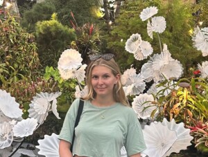 Photograph of the upper body of a woman standing in front of an outdoor backdrop with real and artificial tropical foliage and flowers.