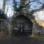A photograph of a man sitting on a bench with a drystone alcove with a rural landscape beyond,