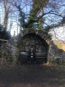 A photograph of a man sitting on a bench with a drystone alcove with a rural landscape beyond,