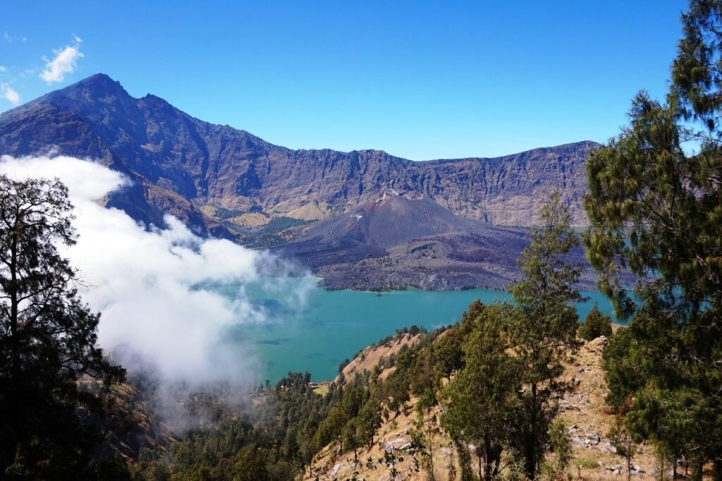 A turquoise volcanic crater lake surrounded by steep rocky cliffs and lush green vegetation, with white clouds partially covering the landscape under a clear blue sky.