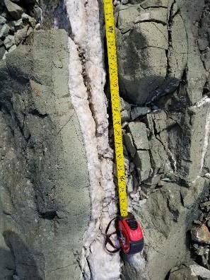 A mineral vein appearing milky white next to a tape measure. The vein is surrounded by altered basalt appearing grey with a green hue. 