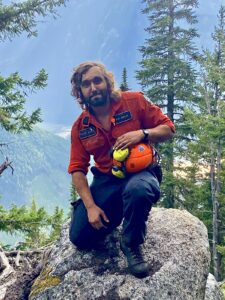 Photo of a man kneeling on a rock in front of a wooded mountainous landscape.