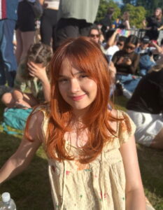 A photo of the head and shoulders of a woman, seated in front of a group of people in an outdoor environment.