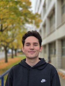 Photo of the head and shoulders of a man standing in front of trees and a building.