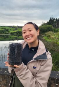 Photograph of a woman holding a glass container of blackberries with a small lake or reservoir in the background.