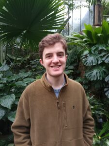 A photo of the upper half of a man standing in front of a background of large tropical leaves.