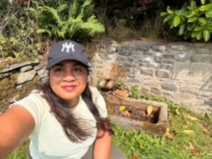 Photo of a crouching woman with a wall and plants in the background.
