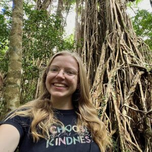 Photo of the head and shoulders of a woman standing in front of tropical trees.