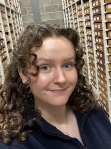 A photo of the head and shoulders of a woman standing in front of archive shelving.