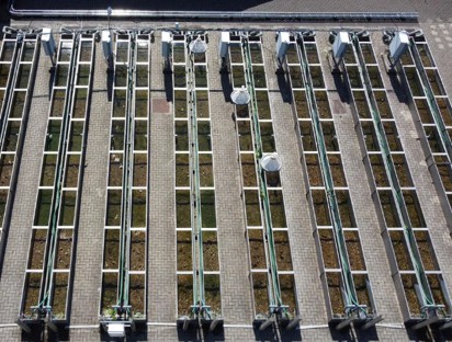 Aerial view of a rectangular wastewater treatment facility containing multiple long, parallel treatment channels arranged in rows. Each channel is separated by paved walkways and fitted with metal rails, support structures, and mechanical equipment positioned along their length. The channels contain water and biological growth of varying colours, ranging from green to brown, indicating different treatment stages or conditions. Several circular and box-shaped control units are mounted above the channels. The image is taken from directly overhead, highlighting the highly ordered, symmetrical layout of the treatment system and its engineered infrastructure.