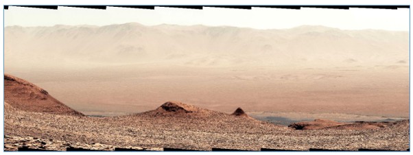 Panoramic view of a barren, rocky landscape with reddish-brown terrain in the foreground and pale, dusty hills stretching across the horizon. The scene is dry and sparsely textured, with no visible vegetation or water, giving the appearance of an arid desert or Mars-like environment.