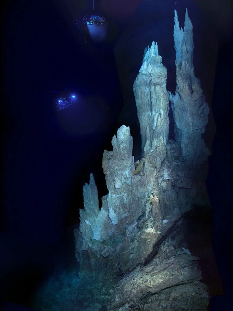A large white/light brown structure with a rough surface is in the foreground. It has a broad base at the bottom of the image with four columns extending upwards to the top of the image. The shortest column is in the centre of the image, with each column increasing in height until the tallest, which is found on the right of the image. In the background, there are two small robotic vehicles that are rectangular prisms. They have lights, and one has a visible tether.