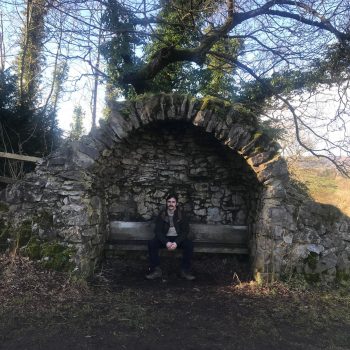 A photograph of a man sitting on a bench with a drystone alcove with a rural landscape beyond,