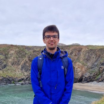 Photograph of a man standing in front of a coastal landscape, with sloping cliffs and a small sandy beach.