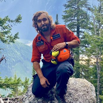 Photo of a man kneeling on a rock in front of a wooded mountainous landscape.