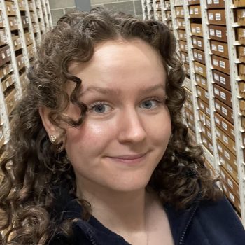 A photo of the head and shoulders of a woman standing in front of archive shelving.