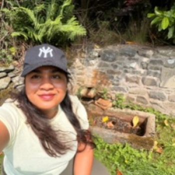 Photo of a crouching woman with a wall and plants in the background.
