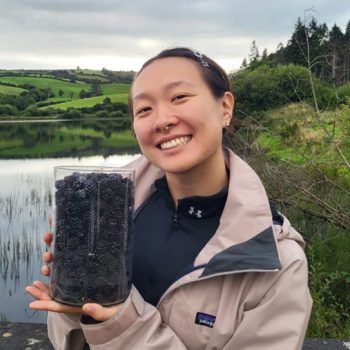 Photograph of a woman holding a glass container of blackberries with a small lake or reservoir in the background.