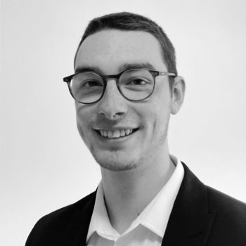 Black and white photograph of the head and shoulders of a man wearing glasses, shirt and jacket.