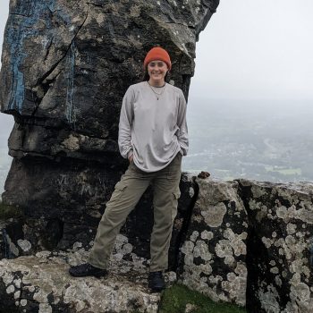 A photograph of a woman standing in front of a tall rocky outcrop.