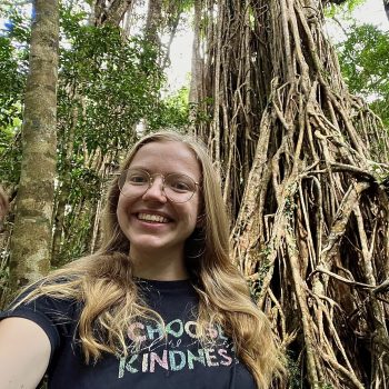Photo of the head and shoulders of a woman standing in front of tropical trees.