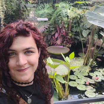 Photo of the head and shoulders of a woman standing in front of a pond with tropical vegetation.