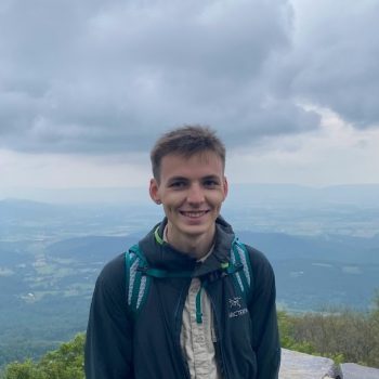 Photograph of a man standing on a high outdoor viewpoint in front of a wide landscape.