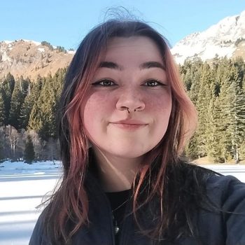 Photograph of the head and shoulders of a woman standing in front of a snowy and forested mountainous background.