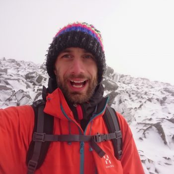 A person in a bright orange jacket and colourful knitted hat with a backpack, standing in front of a snowy mountain landscape.