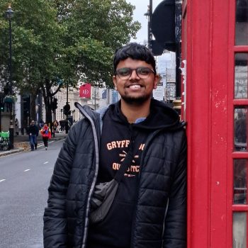 Photograph of a man learning against a traditional red telephone box on an urban street.