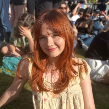 A photo of the head and shoulders of a woman, seated in front of a group of people in an outdoor environment.