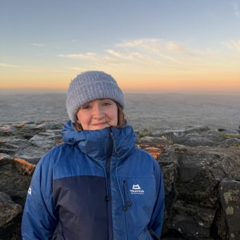 A photograph of the upper body of a woman standing on a rocky outcrop in front of a wide landscape.